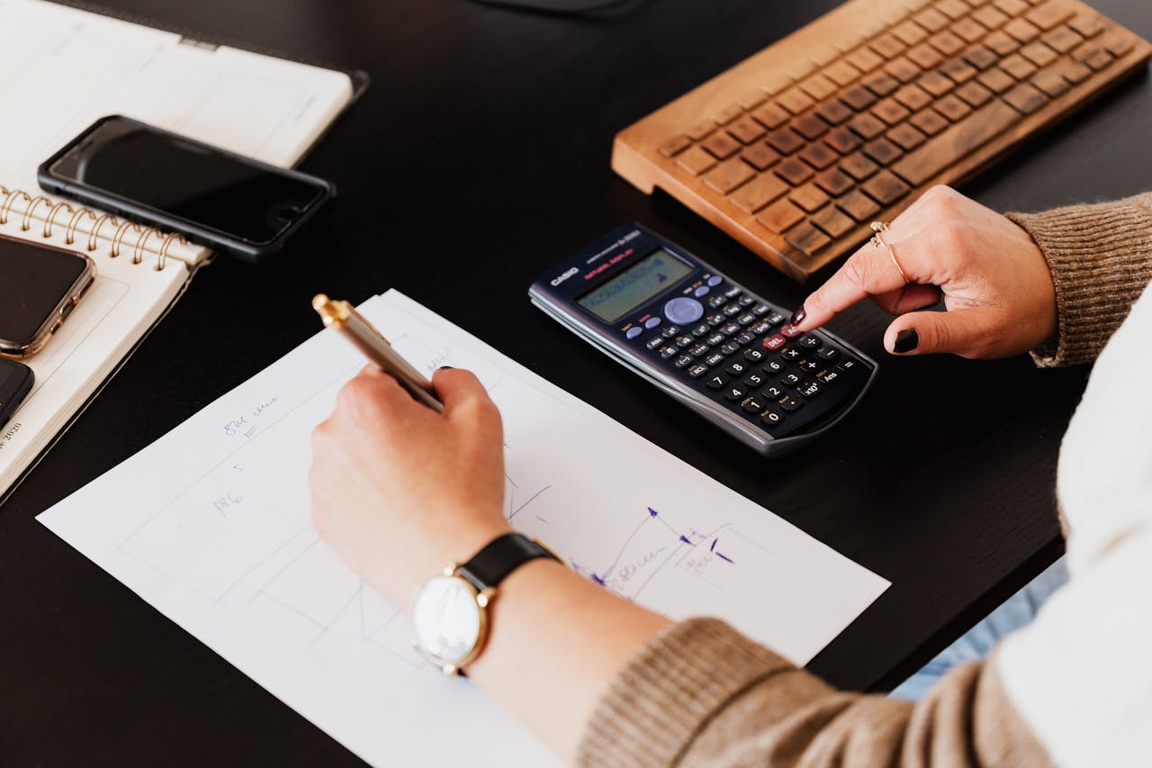 A person calculates with a calculator while writing notes on paper, focused on their work at a desk.